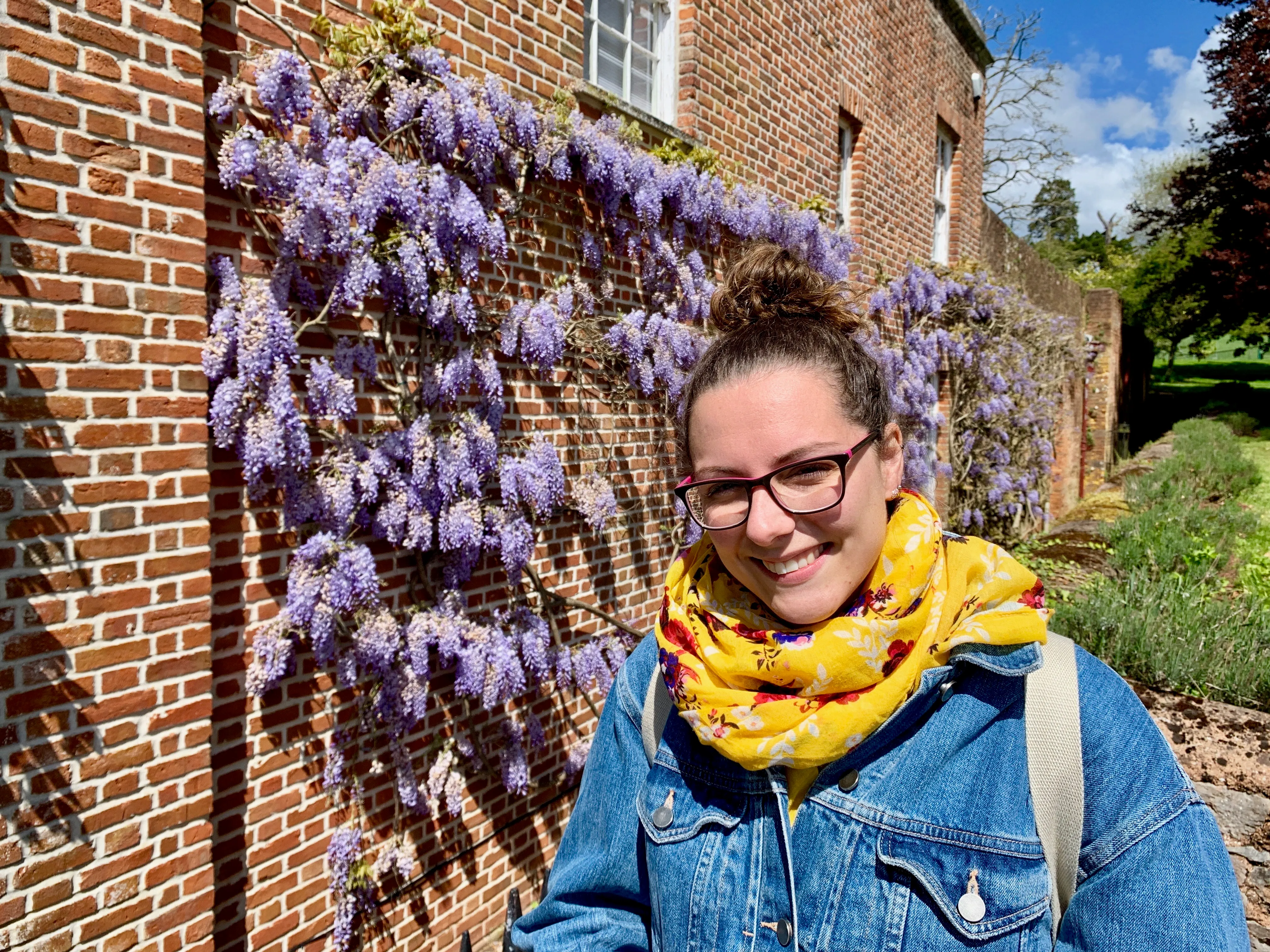 Kirsty smiling in a yellow scarf and denim jacket beside a wisteria-covered brick wall